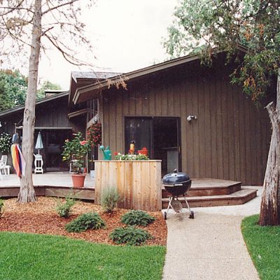 A brown, single-story house with a wooden deck featuring patio furniture and an umbrella, surrounded by trees, green grass, and beautifully maintained bushes by Cordes Family Landscaping and Snow LLC. A charcoal grill sits near the walkway.
