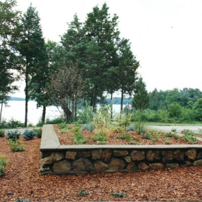A raised stone garden bed with mulch and various plants sits near a road, surrounded by tall trees. Expertly maintained by Cordes Family Landscaping and Snow LLC, the peaceful scene overlooks a body of water and lush greenery.