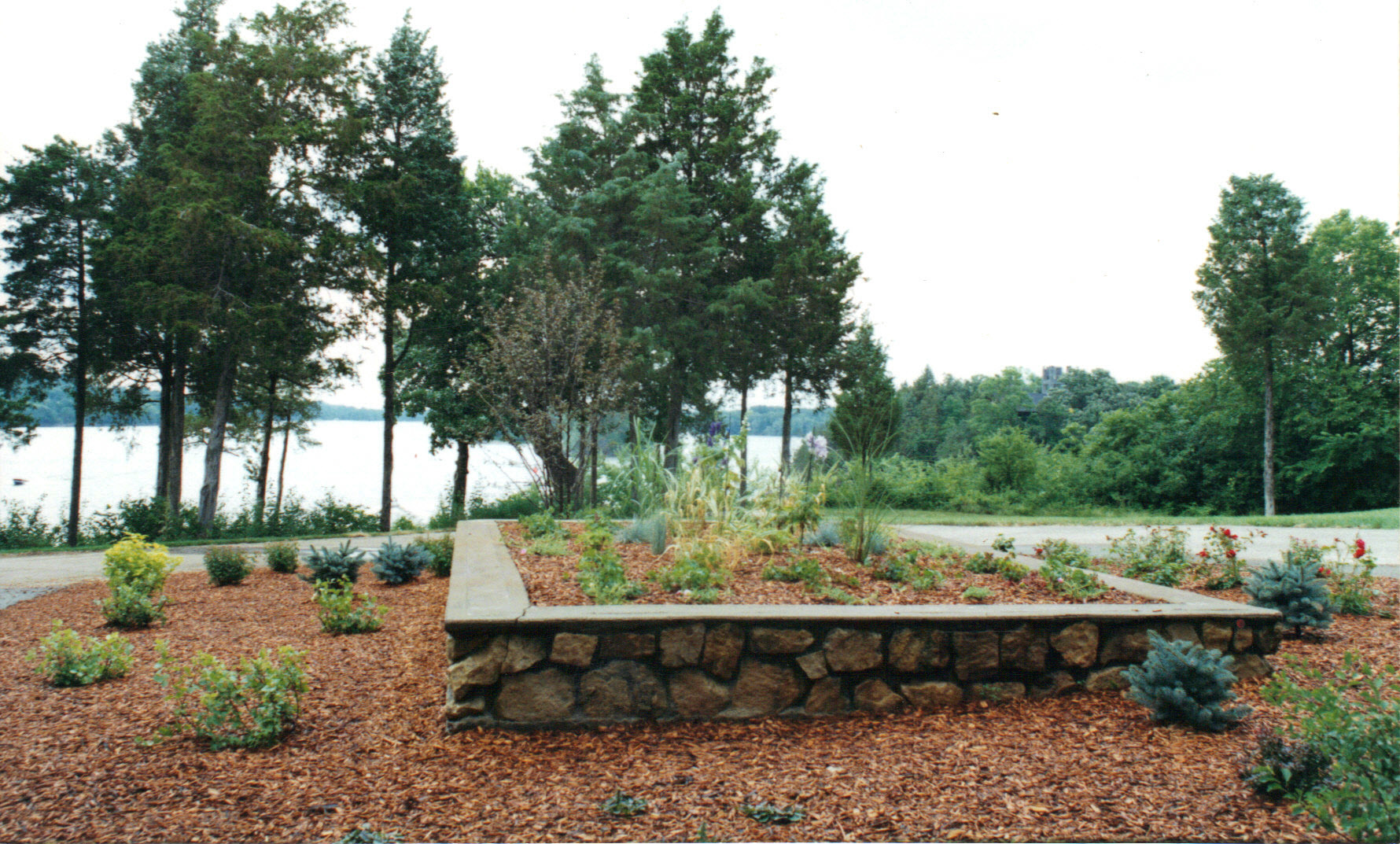 A raised stone garden bed with mulch and various plants sits near a road, surrounded by tall trees. Expertly maintained by Cordes Family Landscaping and Snow LLC, the peaceful scene overlooks a body of water and lush greenery.