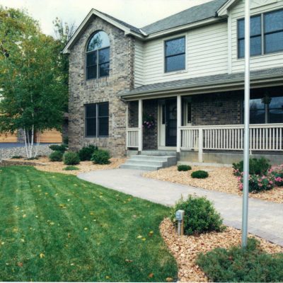 A two-story suburban house with brick and siding exterior, large front porch, and well-maintained landscaping by Cordes Family Landscaping and Snow LLC featuring green lawn, shrubs, flowers, and a stone walkway leading to the entrance.