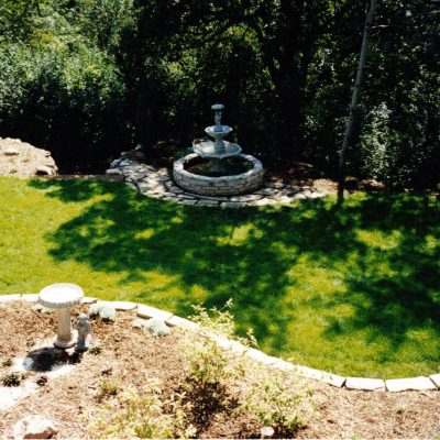 A landscaped backyard by Cordes Family Landscaping and Snow LLC features a circular stone fountain on a grassy lawn, surrounded by trees and mulch beds. A birdbath sits near the foreground on the edge of a curved planting area.