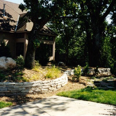 A landscaped yard by Cordes Family Landscaping and Snow LLC features stone retaining walls, large rocks, and mature trees beside a house. A concrete pathway curves through grass and mulch, with sunlight filtering through the trees.