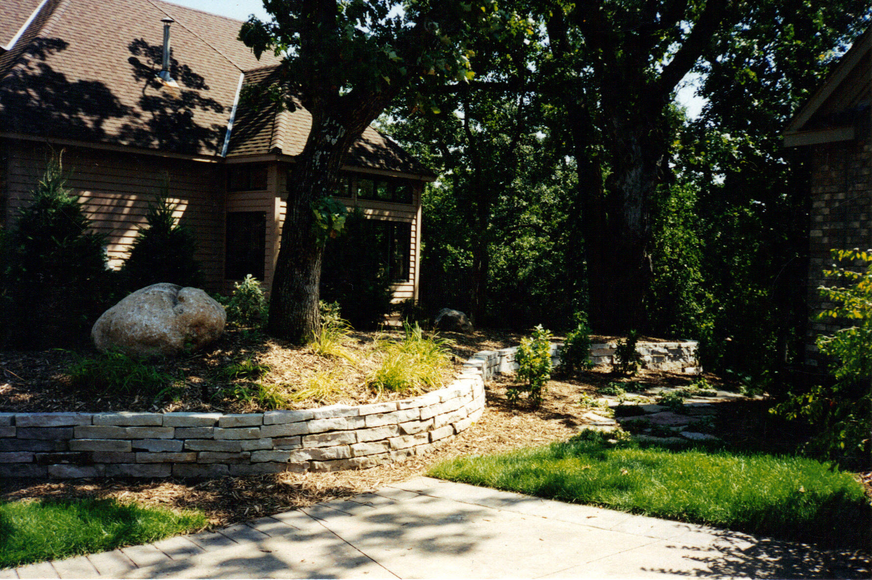 A landscaped yard by Cordes Family Landscaping and Snow LLC features stone retaining walls, large rocks, and mature trees beside a house. A concrete pathway curves through grass and mulch, with sunlight filtering through the trees.