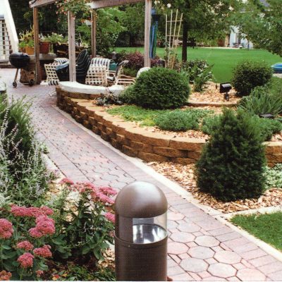 A landscaped backyard features a paved walkway, flower beds, trimmed shrubs, a stone retaining wall, outdoor seating under a pergola, and a garden light in the foreground.