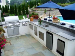 Outdoor kitchen with a built-in grill, stainless steel appliances, sink, and granite countertops next to a swimming pool. There are potted plants, barstools, and large blue umbrellas providing shade. - Cordes Family Landscape & Snow LLC Outdoor kitchen with a built-in grill, stainless steel appliances, sink, and granite countertops next to a swimming pool. There are potted plants, barstools, and large blue umbrellas providing shade.