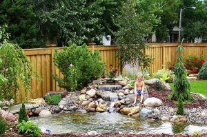 A young child sits by a small backyard pond with rocks and a waterfall, surrounded by lush greenery, trees, and a wooden fence. - Cordes Family Landscape & Snow LLC A young child sits by a small backyard pond with rocks and a waterfall, surrounded by lush greenery, trees, and a wooden fence.