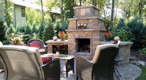 A cozy outdoor patio with wicker chairs, red cushions, a glass table, and a large stone fireplace, surrounded by lush green trees and plants on a sunny day. - Cordes Family Landscape & Snow LLC A cozy outdoor patio with wicker chairs, red cushions, a glass table, and a large stone fireplace, surrounded by lush green trees and plants on a sunny day.