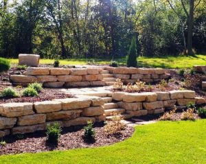 Tiered stone retaining walls with steps, landscaped with mulch, small shrubs, and green grass, set against a backdrop of trees and sunlight.
