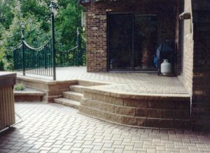 A brick patio with curved steps leads up to a higher level deck area with a railing. There is a sliding glass door, a propane tank, and trees in the background. - Cordes Family Landscape & Snow LLC A brick patio with curved steps leads up to a higher level deck area with a railing. There is a sliding glass door, a propane tank, and trees in the background.