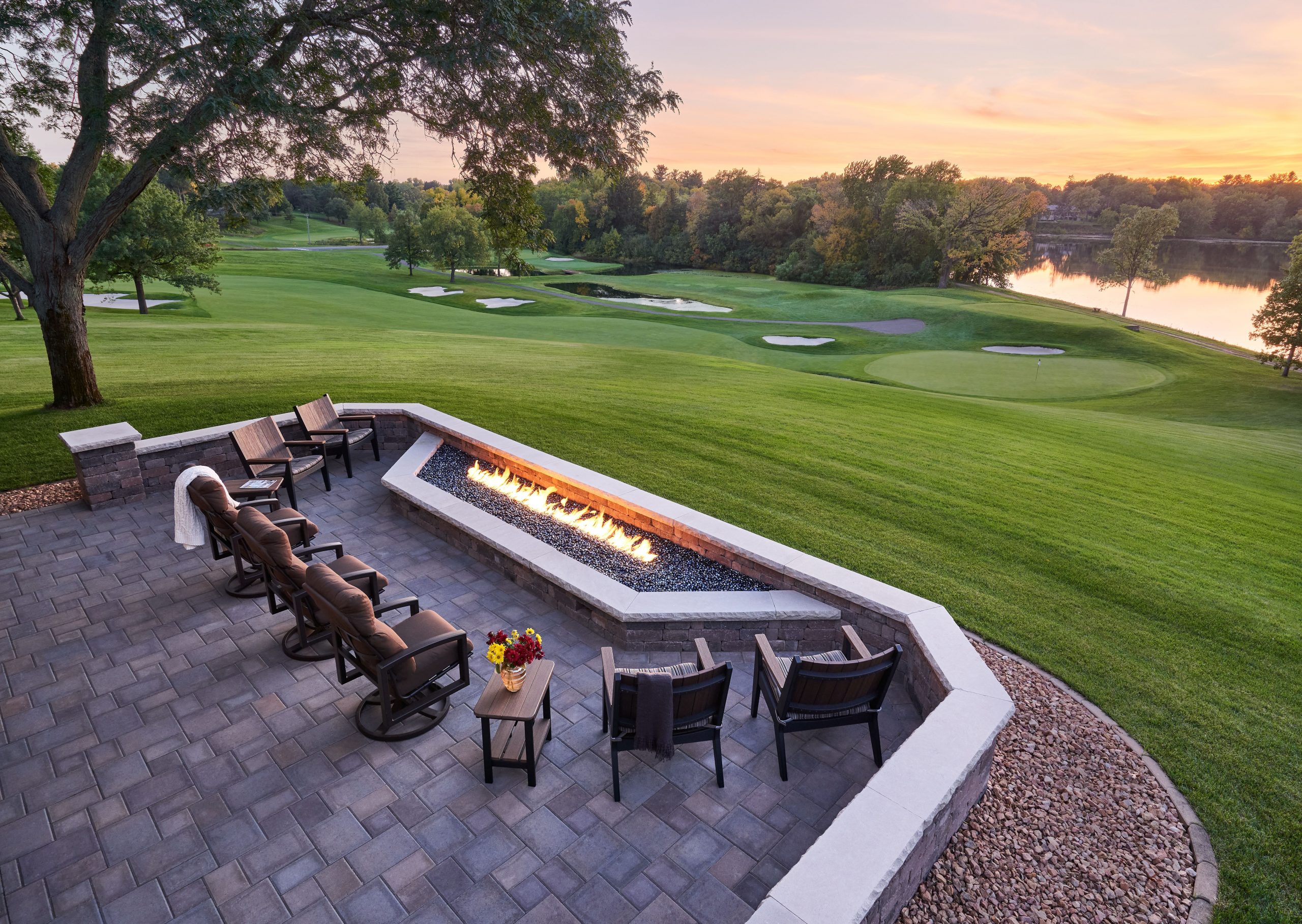 A stone patio with cushioned chairs surrounds a long rectangular fire pit, overlooking a green golf course with trees and a pond at sunset. A small table with flowers sits near the seating area.